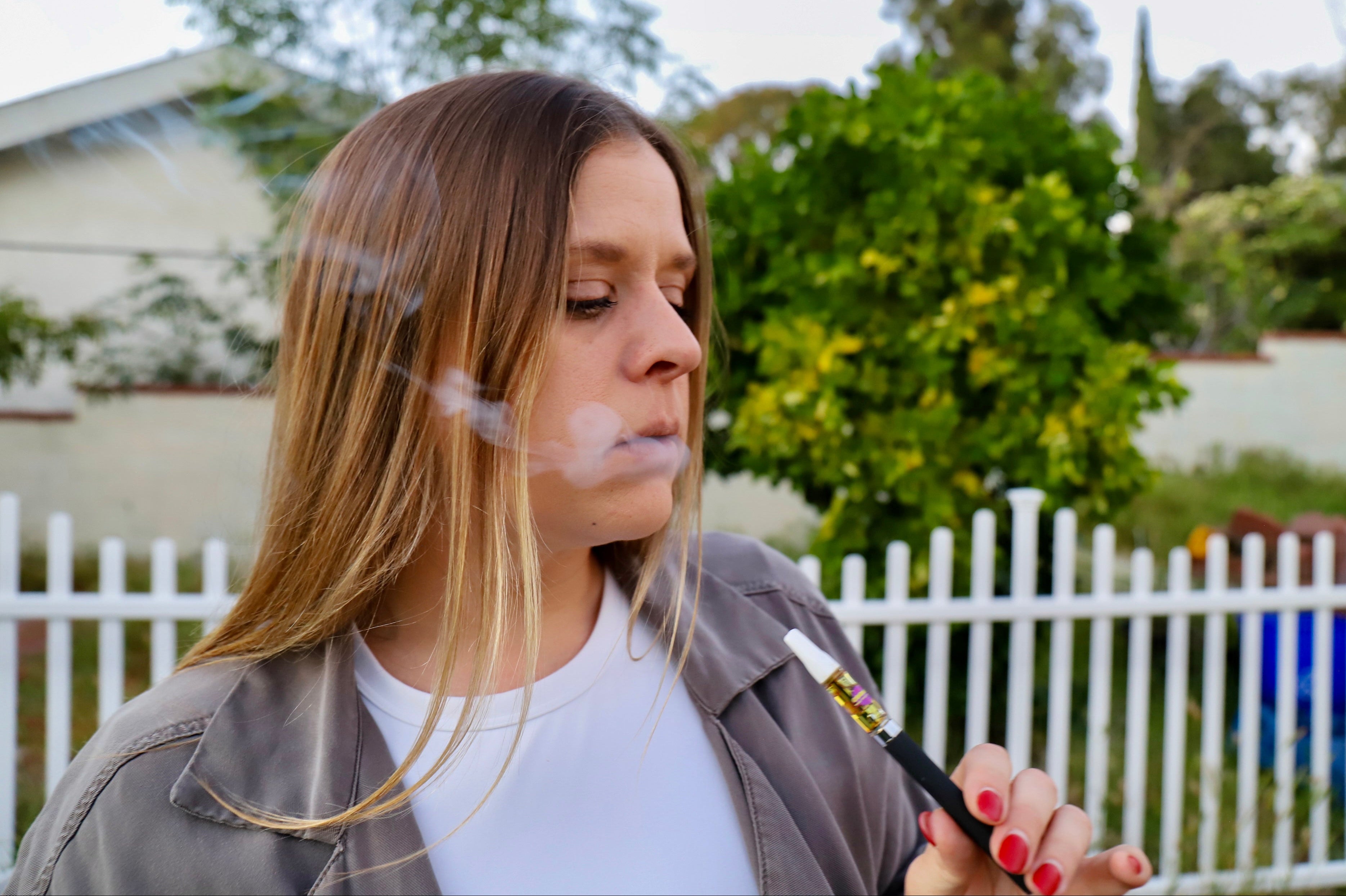 Woman smoking a vape pen outdoors with a white fence and trees in the background