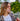 Woman smoking a vape pen outdoors with a white fence and trees in the background